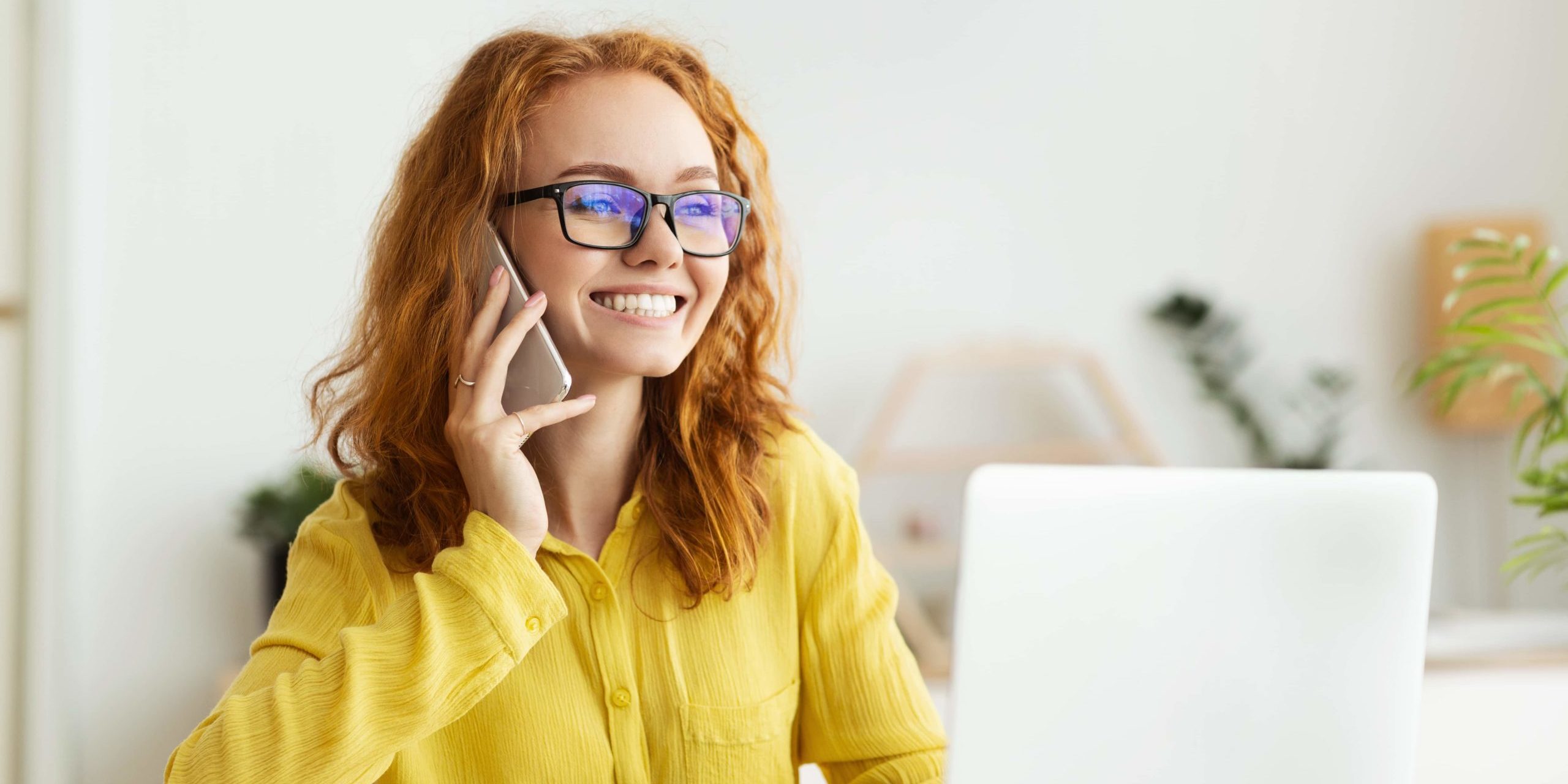 Immagine di una ragazza con occhiali sorridente mentre risponde al cellulare davanti a un computer portatile aperto di fronte a lei